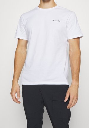 Homme portant un t-shirt blanc uni à manches courtes Columbia et un pantalon noir avec une poche latérale, debout devant un fond uni.