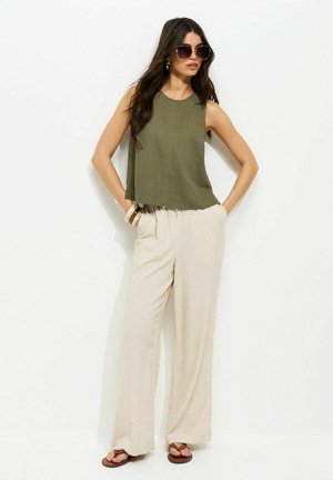 Femme portant de grandes lunettes rondes, un haut sans manches olive, un pantalon beige large, des bracelets et des sandales, posant les mains dans les poches.