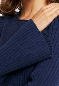 Marinblå ribbad tröja med vida ärmar och rundad halsringning. Texturen verkar mjuk och stickad, med vertikala linjer i tyget.