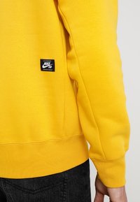 Gul Nike SB sweatshirt med ärm och nederkant på en person som bär svarta jeans, vilket visar det lilla svarta logotypmärket på sidan.