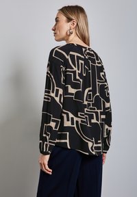 Blusa a maniche lunghe in tessuto nero con un motivo geometrico beige. Dettaglio di pieghe nella parte posteriore, scollatura rotonda e polsini arricciati.