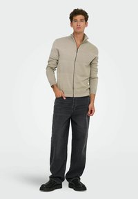 Pull zip beige avec un design texturé ; associé à un jean noir ample et des chaussures noires, se tenant devant un fond clair.