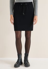 Mujer vistiendo una falda negra hasta la rodilla con cordón, medias negras transparentes, botas negras con cordones y un suéter gris de manga larga con canalé.