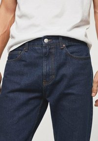 Jeans de mezclilla azul oscuro con un corte clásico, que presentan costuras naranjas, cierre frontal con botón y un diseño tradicional de cinco bolsillos.