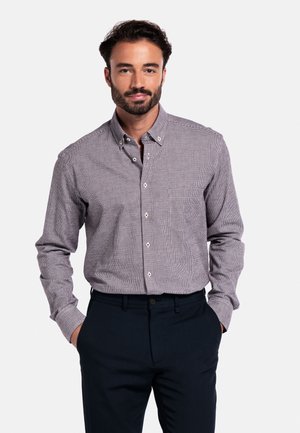 Man met donker haar en een baard, gekleed in een grijs, geblokt overhemd met lange mouwen en donkere broeken, met de handen in de zakken, tegen een witte achtergrond.