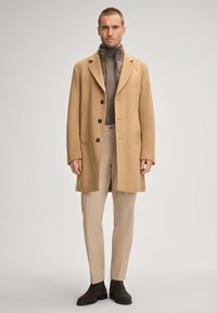 Cappotto di lana beige con bottoni neri, due tasche frontali, sovrapposto a un dolcevita marrone e pantaloni beige chiaro, completato da stivali scuri.