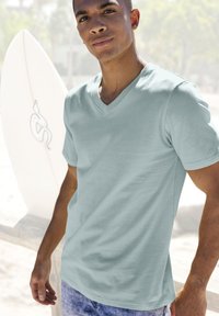 Hellblaues V-Ausschnitt-T-Shirt aus weichem Baumwollmaterial, kombiniert mit hellen Jeansshorts, steht neben einem Surfbrett am Strand.