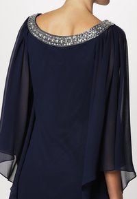 Námořnické modré šaty s průhlednými, splývavými rukávy a zdobeným, klenotem bohatým výstřihem. Měkká textura a elegantní silueta zdůrazňují design.