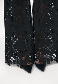 Jambes portant des talons pointus noirs visibles à travers un pantalon noir en dentelle florale avec des motifs découpés complexes.