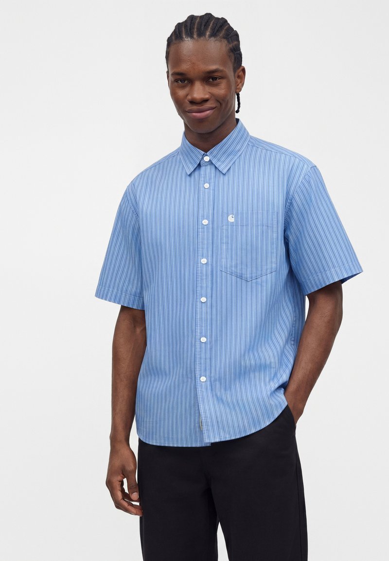 Man with braided hair wearing a light blue, short-sleeve, button-up shirt with thin stripes and black pants against a plain background.