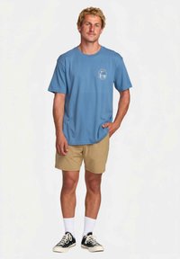 Camiseta de algodón azul claro con mangas cortas y un gráfico en el pecho, combinada con pantalones cortos caqui. Zapatillas con cordones blancos y calcetines tipo crew.