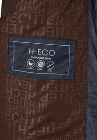 Marinefarbendes Etikett auf braunem Textil mit wiederholendem "HECHTER"-Text, das "H-ECO Sustainable Fabric" zeigt sowie Symbole für nachhaltig, umweltfreundlich und wasserabweisend.