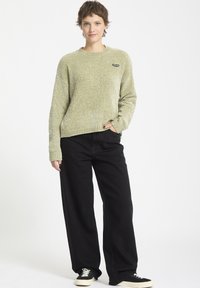 Pull en mailles de couleur vert clair avec un col rond et un petit logo noir, associé à un pantalon noir à jambes larges et à des baskets noires.