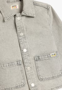 Chaqueta de mezclilla gris con cuello, botones frontales, dos bolsillos en el pecho y una etiqueta amarilla "NYC SK8" en el bolsillo derecho.