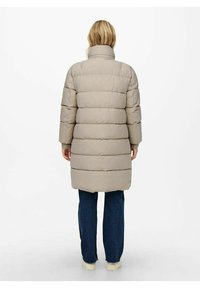 Kvinde iført en knælang beige pufferjakke, blå jeans og hvide sneakers, som står med ryggen til mod en ensfarvet lys baggrund.