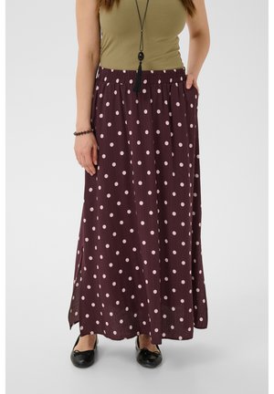 Femme portant une longue jupe violette à pois rose clair, un débardeur vert olive, des ballerines noires et un long collier à pampilles noires.
