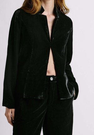 Femme portant une veste et un pantalon en velours noir, veste partiellement ouverte révélant le ventre nu, une main dans la poche.