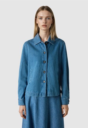 Jonge vrouw met steil blond haar, gekleed in een blauwe denimjas met knopen en een bijpassende rok, staand tegen een effen lichte achtergrond.