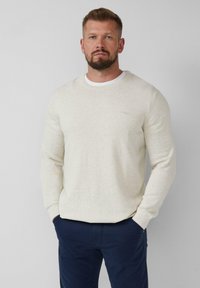 Mann mit Bart, der einen cremefarbenen Strickpullover über einem weißen Hemd und einer dunkelblauen Hose trägt, steht mit den Händen in den Taschen vor einem schlichten Hintergrund.