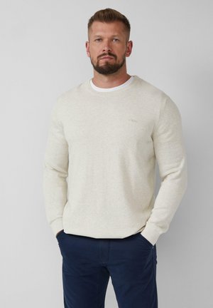 Homme avec une barbe portant un pull en tricot crème sur une chemise blanche et un pantalon bleu marine, se tenant avec les mains dans les poches devant un fond uni.