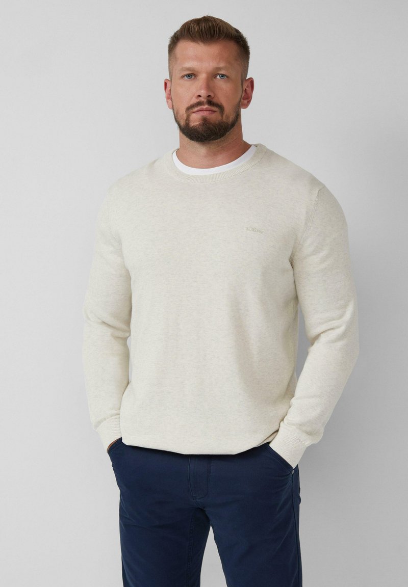 Mann mit Bart, der einen cremefarbenen Strickpullover über einem weißen Hemd und einer dunkelblauen Hose trägt, steht mit den Händen in den Taschen vor einem schlichten Hintergrund.