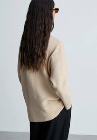 Maglione di lana beige con vestibilità rilassata, orlo a coste e dettagli ai polsi. Indossato con pantaloni neri, con capelli lunghi e mossi dietro.