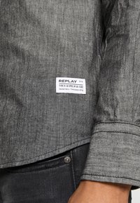Cuff di una camicia grigia a maniche lunghe con un'etichetta bianca "Replay" sopra la tasca dei jeans neri e un dettaglio con bottone sul braccio di una persona.