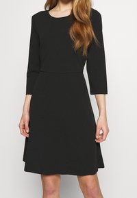 Czarna sukienka z długim rękawem, okrągłym dekoltem i dopasowaną talią, delikatnie rozszerzająca się na dole. Wykonana z gładkiego, teksturowanego materiału.