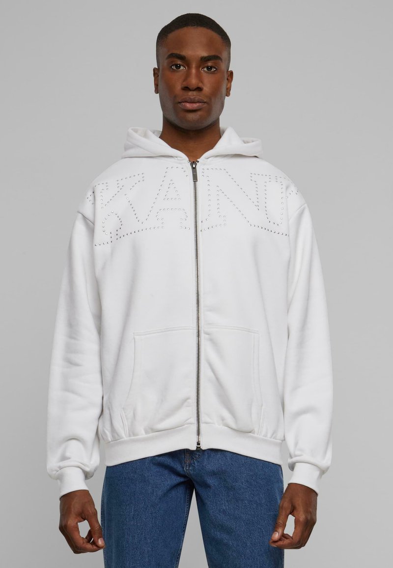 Hvit glidelås hettegenser med et stort "KANI" design i nagler, med hette, frontlommer og ribbede mansjetter, brukt over blå jeans.