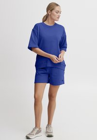 Blå bomulds t-shirt med korte ærmer og løs pasform, parret med matchende blå shorts. Hvide og grå sneakers fuldender outfittet.