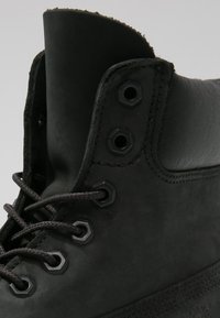 Des bottes en cuir noir présentent une pointe ronde, une semelle en caoutchouc, des œillets en métal et des lacets noirs. La tige a une texture lisse avec un soutien renforcé de la cheville.