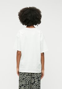 Χαλαρή λευκή βαμβακερή μπλούζα με στρογγυλή λαιμόκοψη και κοντά μανίκια, συνδυασμένη με μαύρο παντελόνι με γεωμετρικά σχέδια.