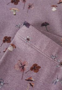 Zbliżenie na fioletową tkaninę z nadrukowanymi małymi kwiatami i grzybami w brązowych, pomarańczowych i białych tonach, ukazujące złożony szew.