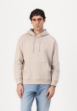 Mann trägt beige Hoodie mit Fronttasche und hellblaue Jeans, steht vor einfarbigem weißem Hintergrund.