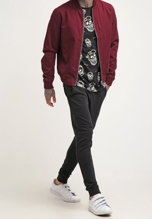 Chaqueta bomber granate sobre una camiseta negra con gráficos de calaveras blancas, combinada con pantalones jogger negros y zapatillas blancas. Tatuajes visibles en los brazos.