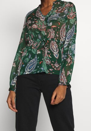 Femme portant un chemisier vert à manches longues avec un motif cachemire multicolore et un pantalon noir, debout devant un fond uni.
