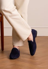 Persoon in beige broek en donkerblauwe suède instap schoenen, zittend met gekruiste benen op een houten vloer naast een stoel.