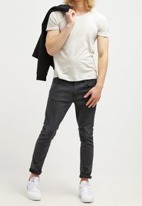 Harmaa istuva t-paita, tummanharmaat slim-fit farkut, valkoiset tennarit. Malli pitää mustaa takkia. Sujuva materiaali, minimalistinen muotoilu.