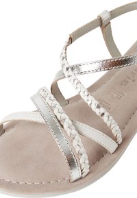 Sandal med stropper, der har flettede hvide og skinnende sølvfarvede remme, en blød beige ruskindsindersål og en glat hvid ydersål.