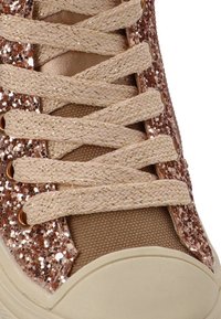 Zapatilla con un cuerpo de tela color marrón claro, lentejuelas doradas rosadas brillantes en la parte superior, y cordones beige claros entrelazados a través de ojales de metal.