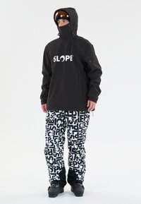 Sort hættejakke med stort hvidt "SLOPE"-tryk, parret med hvide bukser med et dristigt sort grafisk mønster. Begge dele er designet til vintersport.