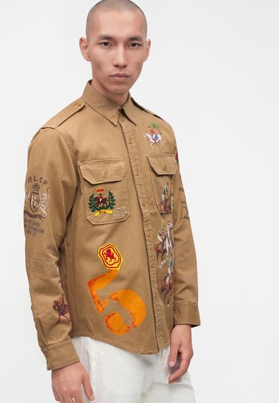 Mann trägt ein hellbraunes Hemd mit Knöpfen, bestickt mit Reitermotiven, Wappen und orangefarbenen abstrakten Mustern, kombiniert mit weißen Hosen, vor weißem Hintergrund.