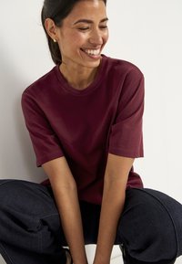 Glunderende vrouw met gouden oorringen, gekleed in een bordeauxkleurig shirt met korte mouwen en donkere broek, zittend met haar armen op haar knieën tegen een witte muur.