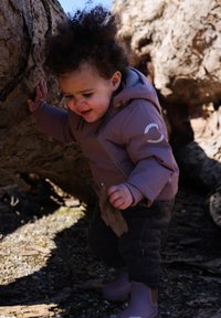 Veste à fermeture éclair de couleur violet clair avec capuche ; pantalon gris à motifs ; bottes en caoutchouc ; enfant tenant un morceau d'écorce près d'un tronc d'arbre.