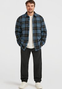 Mand iført en blå og sort ternet skjorte over en hvid t-shirt, sorte bukser og hvide sneakers, står med hænderne i lommerne.