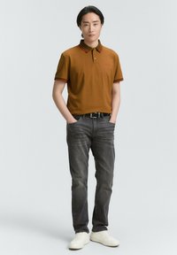 Camisa polo marrón con acentos rojos, combinada con jeans gris oscuro y zapatillas blancas. La camisa tiene cuello y mangas cortas.