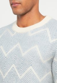 Jasnoniebieski sweter wykonany z miękkiej dzianiny, przedstawiający wzór w zygzaki. Biała, prążkowana okrągła szyja. Tekstura wydaje się puszysta i przytulna.
