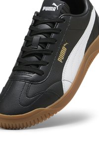 Schwarze Puma Sportschuhe mit weißem Streifen, Gummisohle, schwarzen Schnürsenkeln und goldenem Puma-Logo an der Seite sowie weißem Puma-Logo auf der Zunge.