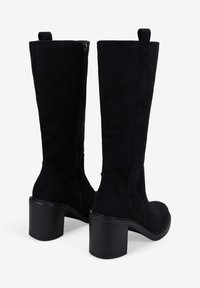 Bottes montantes en suède noir avec un talon carré, dotées d'une fermeture éclair sur le côté et d'une languette à l'arrière. Texture lisse et design épuré.