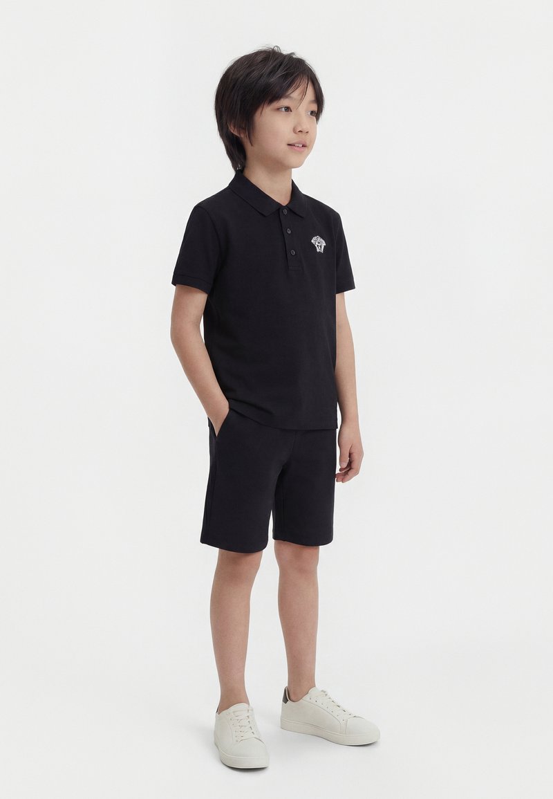 Αγόρι που στέκεται με τα χέρια στις τσέπες, φορώντας μαύρο πόλο μπλουζάκι, μαύρα σορτς και λευκά αθλητικά παπούτσια, στο υπόβαθρο απλού λευκού.